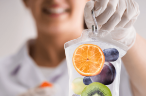 Eine medizinische Fachkraft mit Handschuhen hält einen Infusionsbeutel, der mit frischem Obst wie Orange, Kiwi, Traube und Pflaume gefüllt ist – symbolisch für eine vitaminreiche Infusion.