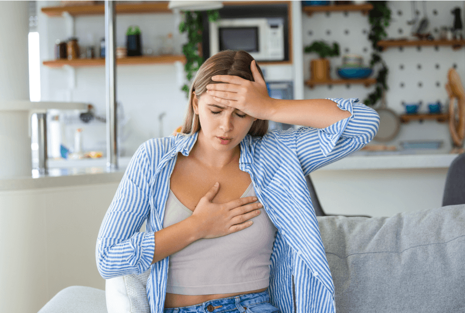 Junge Frau hält sich die Brust und den Kopf, zeigt Symptome von Unwohlsein und Schwindel, in einem modern eingerichteten Wohnzimmer – Konzept von gesundheitlichen Beschwerden oder Stress im Alltag.