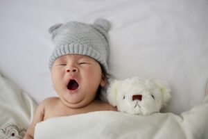 Gaehnendes Baby mit Muetze und Teddybaer im Bett, Symbolbild fuer die 6-Monats-Schlafregression