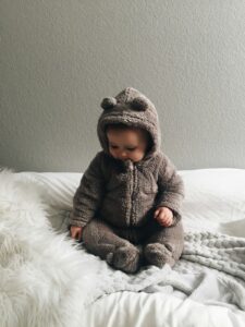 Baby im Kuschel-Strampler sitzt aufrecht auf dem Bett, Symbolbild f&uuml;r den 8-Monats-Schlafschub mit neuen motorischen Meilensteinen
