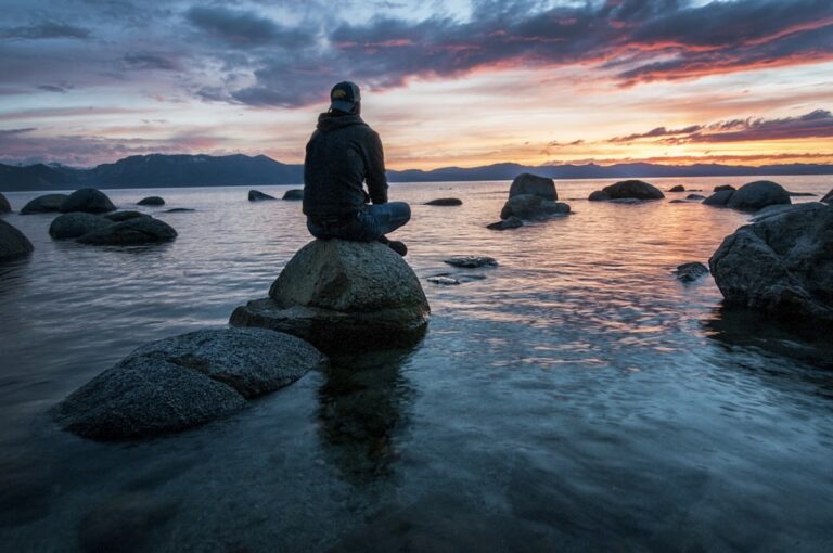Person sitzt meditierend auf einem Felsen am Wasser bei Sonnenuntergang, Symbolbild fuer die Wim-Hof-Atemtechnik