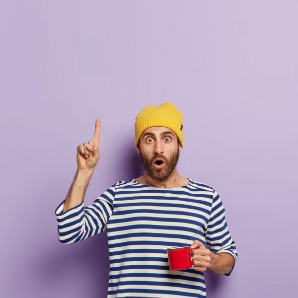 impressed-millennial-guy-points-upwards-with-index-finger-has-shocked-expression-drinks-coffee-morning-holds-red-mug-wears-yellow-hat-striped-sailor-jumper-demonstrates-something-up
