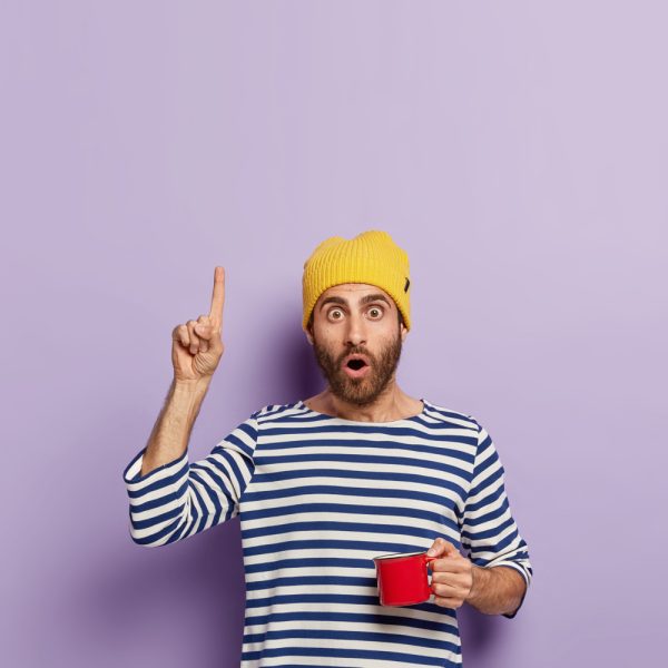 impressed-millennial-guy-points-upwards-with-index-finger-has-shocked-expression-drinks-coffee-morning-holds-red-mug-wears-yellow-hat-striped-sailor-jumper-demonstrates-something-up impressed-millennial-guy-points-upwards-with-index-finger-has-shocked-expression-drinks-coffee-morning-holds-red-mug-wears-yellow-hat-striped-sailor-jumper-demonstrates-something-up